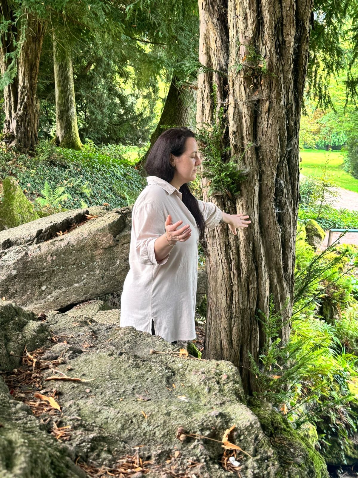 Auf dem Bild sieht man Silvia Lenk, rechts von der Seite. Sie steht in einem Wald. Sie hat ihre linke Hand auf einen dicken Baum gelegt und ihre rechte Hand von sich sanft weggestreckt. Sie trägt eine weiße Bluse, die nach vorne offen ist, drunter trägt sie ein schwarzes T-Shirt. Ihre fast schwarzen, langen Haare trägt sie offen auf ihre linke Seite gelegt. Man sieht im Hintergrund eine grüne Lichtung, mit Bäumen und Büschen. Hinter ihr sind Steine und Baumstämme. Rechts vor ihr liegt ein großer Felsen. Sie hat die Augen geschlossen.