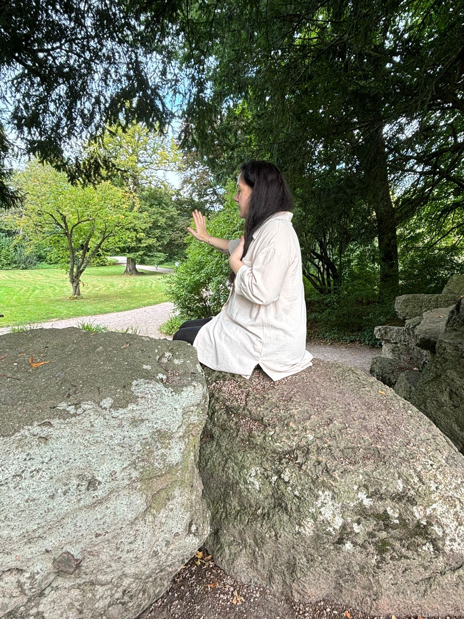 Auf dem Bild sieht man Silvia Lenk, links, schräg hinten von der Seite. Sie sitzt auf einem großen Felsen und neben ihr, auf der linken Seite, steht ein weiterer großer Felsen. Sie sitzt auf diesen Felsen hat die linke Hand auf ihrer Brust, die rechte Hand sanft von sich weggestreckt. Ihre fast schwarzen Haare trägt sie offen und locker auf ihrer linken Schulter. Im Hintergrund sieht man einen Park. Auf ihrer rechten Seite sieht man einen großen Baum und im Hintergrund einen Fußgängerweg, mit schönen Bäumen, einer Wiese und blauen Himmel im Hintergrund. Silvia Link scheint sehr entspannt.